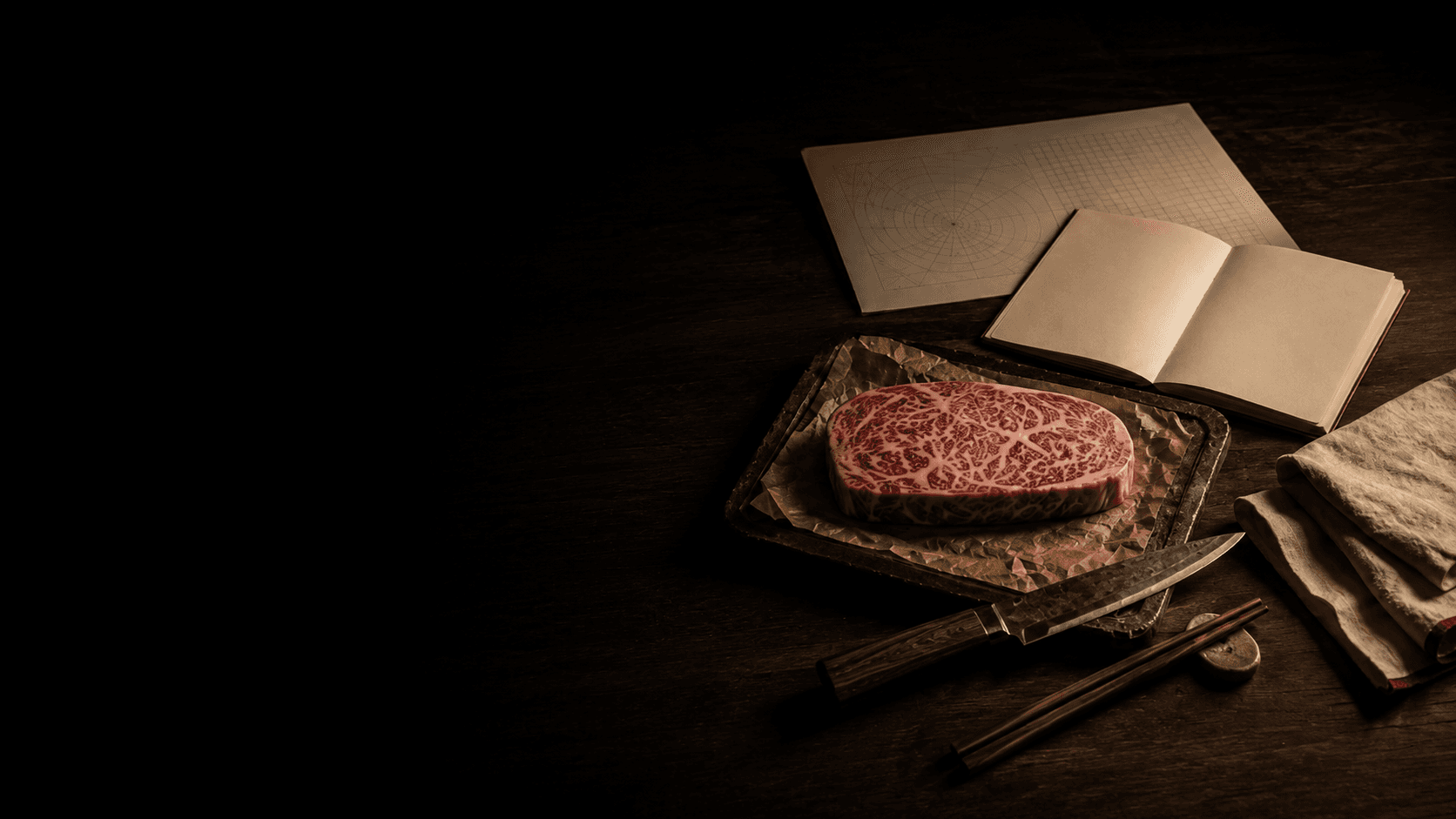 A marbled Wagyu steak on butcher parchment beside a knife, chopsticks, linen and an open notebook on a dark wood table