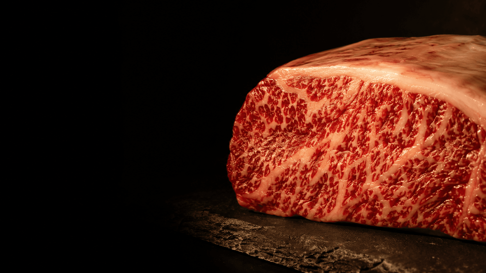 Extreme close-up of a Kobe-grade strip loin, its fine white lattice of marbling glowing under warm gold light on dark slate