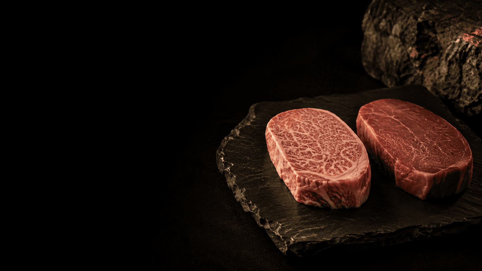 A densely marbled Wagyu steak with a fine white lattice of fat on dark slate, beside a plain lean commodity steak, under dramatic side light